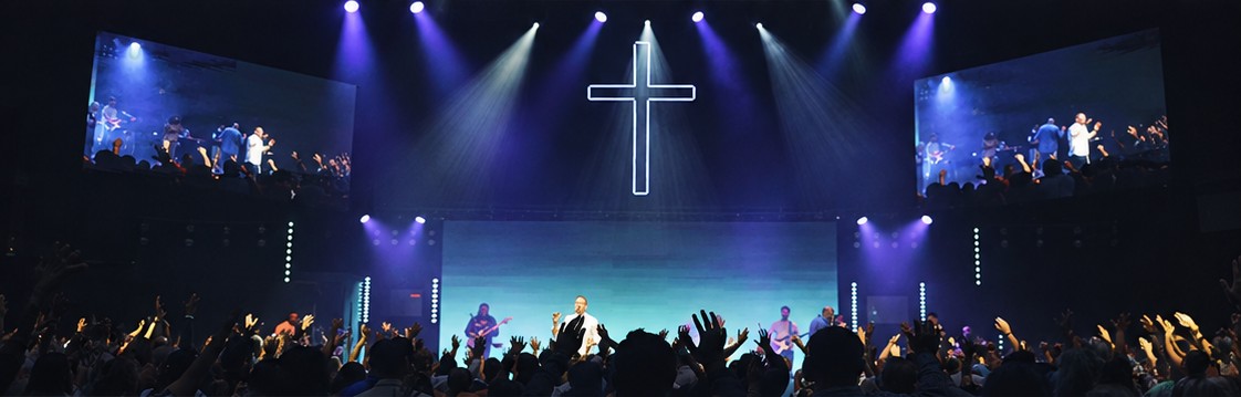 Culto na Church In The Son, com a banda no palco, a cruz iluminada, os telões e o público de mãos levantadas